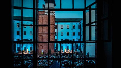 Na pierwszym planie stare, fabryczne okno przez które widać dziedziniec Centralnego Muzeum Włókiennictwa w Łodzi. Na dziedzińcu duża grupa siedzących ludzi zwróconych w stronę sceny, na której grają muzycy.