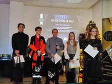 Na zdjęciu widzimy pięć osób stojących w jasnym pomieszczeniu na tle dużego ekranu z białym napisem WYRÓŻNIENIE. Każda z osób w dłoniach trzyma czarne torby i białe kartki A4. Jedna z osób trzyma w ręku mikrofon.