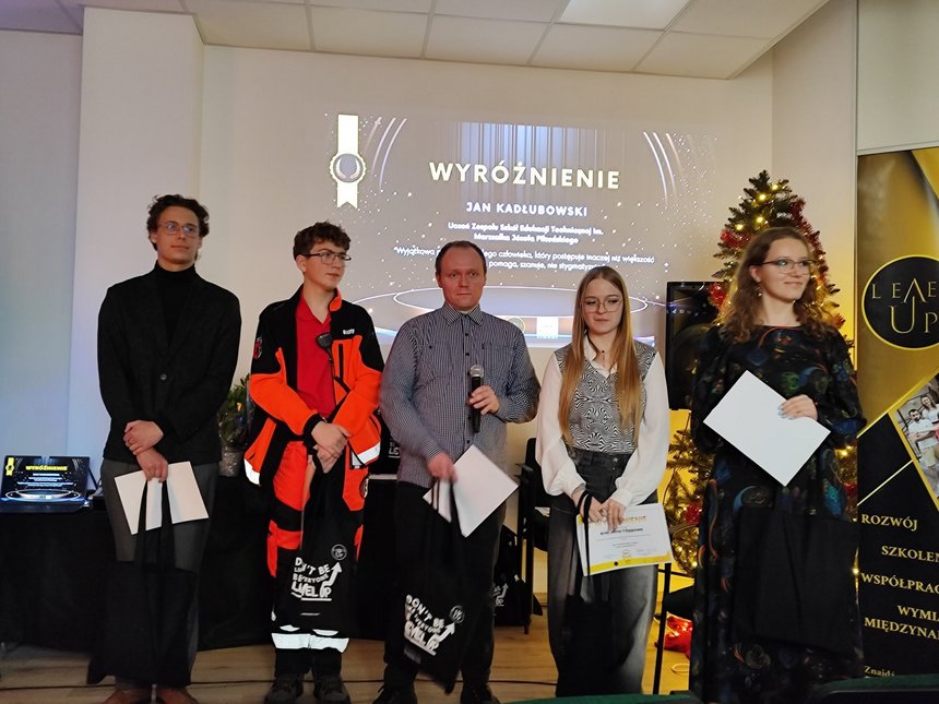 Na zdjęciu widzimy pięć osób stojących w jasnym pomieszczeniu na tle dużego ekranu z białym napisem WYRÓŻNIENIE. Każda z osób w dłoniach trzyma czarne torby i białe kartki A4. Jedna z osób trzyma w ręku mikrofon.