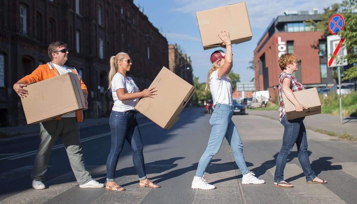 Czwórka Pracowników Oddziału Wsparcia Mieszkańców przechodzi na pasach niosąc w rękach duże kartonowe pudła do przeprowadzek. W tle ceglane budynki famuł przy ulicy Ogrodowej. Kompozycja zdjęcia nawiązuje do słynnej okładki płyty Abbey Road zespołu The Beatles.