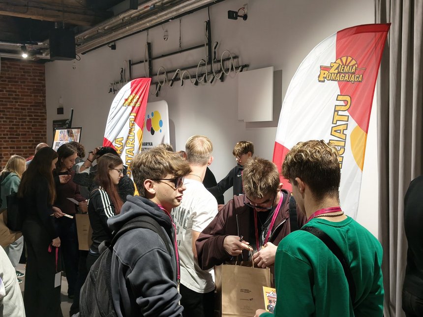 Na zdjęciu widzimy grupy młodzieży po trzy, cztery osoby, które ze sobą rozmawiają. Za nimi widzimy windery w kolorach białym, żółtym, czerwonym z żółtym napisem Ziemia Pomagająca. W tle widzimy białą ścianę z punktami świetlnymi. 