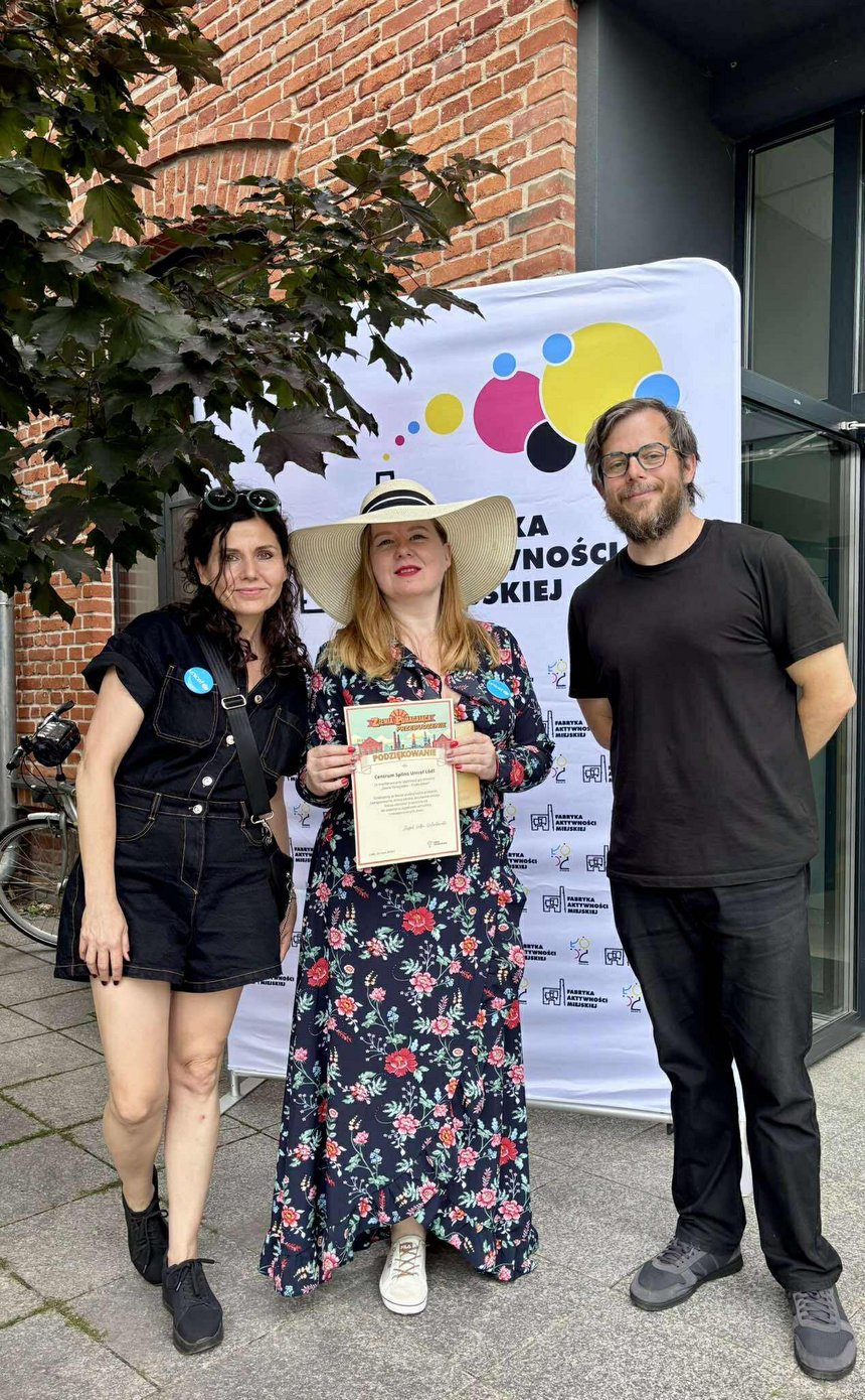 Na zdjęciu widać trzy osoby (jedna z nich ma jacny kapelusz na głowie) na tle kolorowego banneru reklamowego z logo fabryki i napisem Fabryka Aktywności Miejskiej. Jedna z osób trzyma w ręku kartkę A-4 z napisami: Ziemia Pomagająca Przebudzenie. Podziękowanie dla „Spilno” za uczestnictwo w grze miejskiej. W lewym górnym rogu widać fragment korony drzew na tle fragmentu kamienicy z czerwonej cegły.