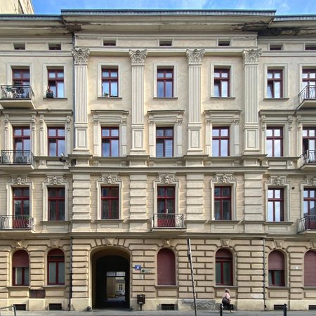 Na zdjęciu znajduje się frontowa elewacja dużej, czterokondygnacyjnej kamienicy. Budynek jest symetryczny i ma jasną, kremowo-beżową fasadę z bogatymi zdobieniami architektonicznymi. Każda kondygnacja ozdobiona jest gzymsami. Całość sprawia wrażenie zadbanego, historycznego budynku miejskiego.