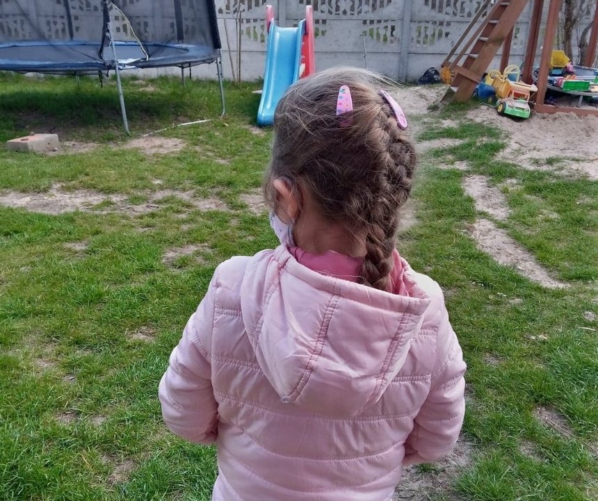 Dziewczynka w różowej kurtce odwrócona plecami stoi pośrodku pustego placu zabaw.