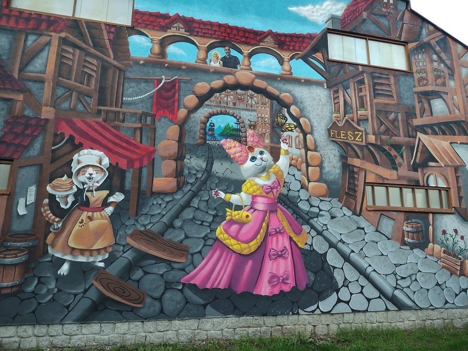 , Kolorowy mural przedstawiający na pierwszym planie dwa koty przebrane w średniowieczne stroje. Biały kot przebrany jest w różową suknię księżniczki z żółtą torebką w kształcie rybki. Drugi kot przebrany za służbę trzymający talerz z naleśnikami w łapce. W tle na drugim planie widać wioskę w średniowiecznym stylu. 