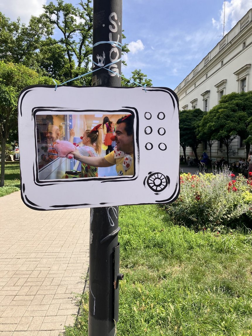 Na zdjęciu widać słup uliczny, do którego przymocowana jest dekoracja – papierowa lub kartonowa rama w kształcie starego telewizora. W jej wnętrzu znajduje się wydrukowane kolorowe zdjęcie przedstawiające o kolorowy korytarz prawdopodobnie w szpitalu i osoby z kolorową piłeczką na nosie. Jedna z osób trzyma w dłoni maskotkę . W tle widzimy trawniki, kwiaty, drzewa i duży budynek.