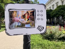 Na zdjęciu widać słup uliczny, do którego przymocowana jest dekoracja – papierowa lub kartonowa rama w kształcie starego telewizora. W jej wnętrzu znajduje się wydrukowane kolorowe zdjęcie przedstawiające o kolorowy korytarz prawdopodobnie w szpitalu i osoby z kolorową piłeczką na nosie. Jedna z osób trzyma w dłoni maskotkę . W tle widzimy trawniki, kwiaty, drzewa i duży budynek.