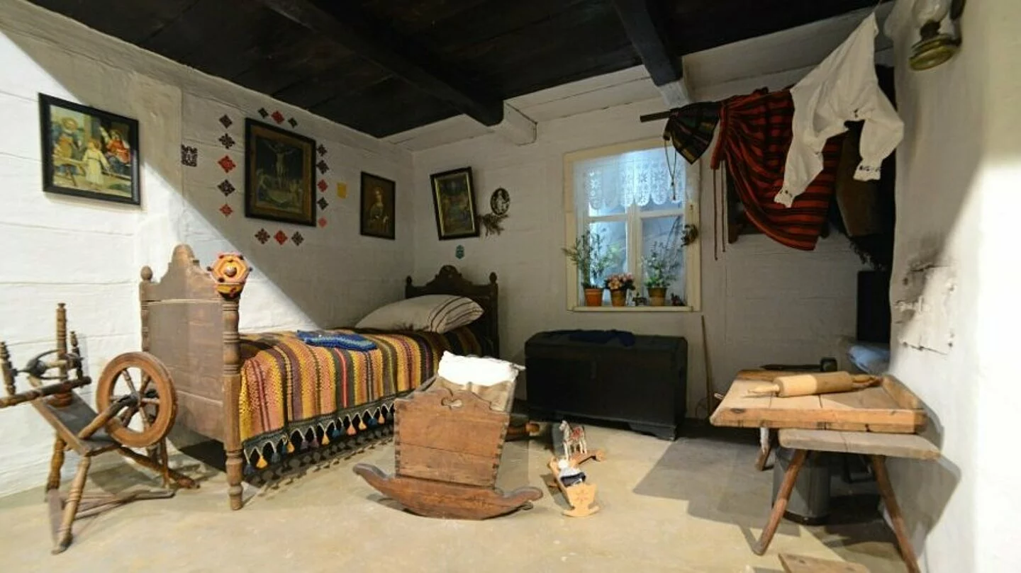  , Na zdjęciu wystawa muzeum - widać historyczny pokój, a w nim stare pojedyncze łóżko w lewym rogu pokoju. Przy łóżku znajduje się maszyna do przędzenia. Na środku pokoju stoi zabytkowa kołyska. W prawym górnym rogu pokoju wiszą ubrania.