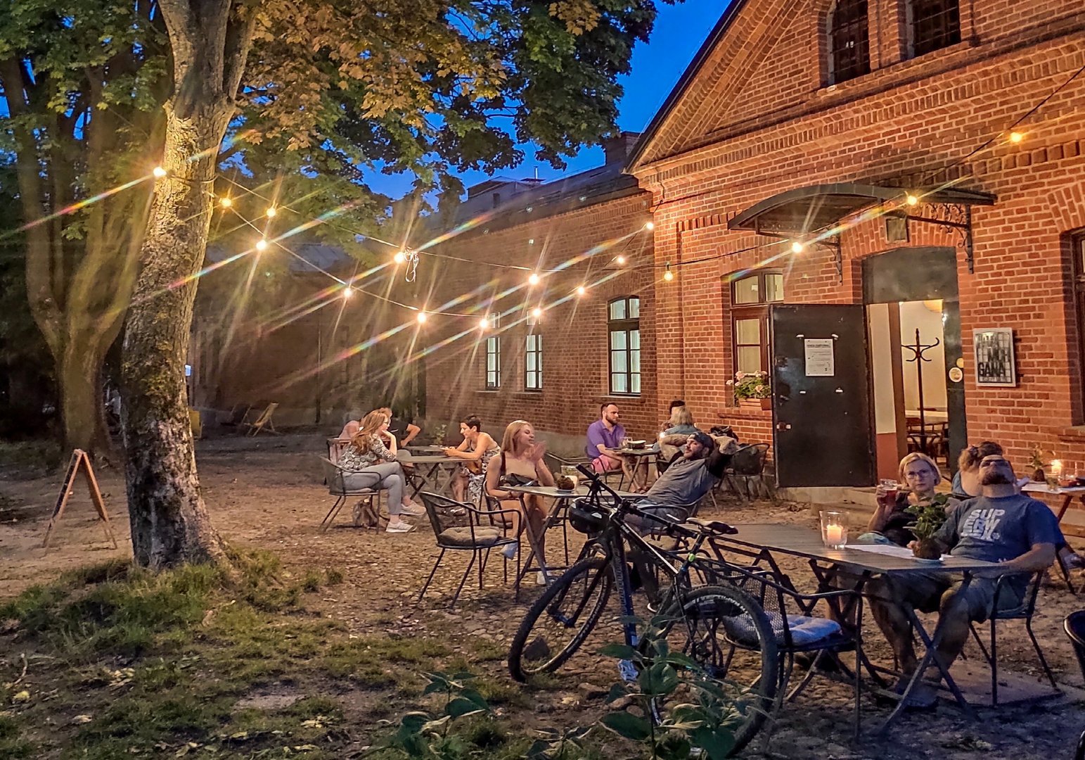Letni wieczór. Ludzie siedzący przy stolikach przed budynkiem z czerwonej cegły. Na pierwszym planie rower postawiony przy stoliku.