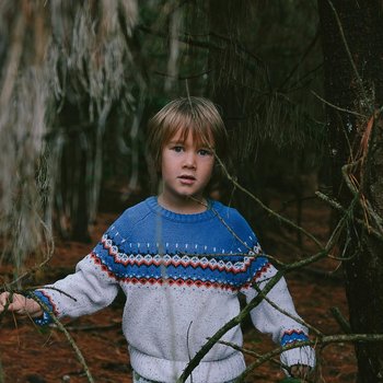 Chłopiec w szaro-niebieskim swetrze stoi pośrodku lasu.