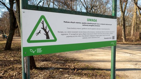 Tablica informująca o zagrożeniu przy wejściu do parku. Na tablicy grafika trójkąt z zielona obwódką, w środku fragment drzewa z odłamującą się gałęzią. Napis Uwaga, podczas silnych wiatrów zachowaj szczególną ostrożność.