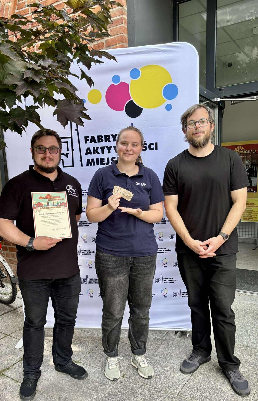 Na zdjęciu widać trzy osoby na tle kolorowego banneru reklamowego z logo fabryki i napisem Fabryka Aktywności Miejskiej. Jedna z osób trzyma w ręku kartkę A-4 z napisami: Ziemia Pomagająca Przebudzenie. Podziękowanie dla „Fundacji Aktywnej Rehabilitacji FAR” za uczestnictwo w grze miejskiej. Druga z osób w ręku trzyma brązowy kartonik. W lewym górnym rogu widać fragment korony drzew.