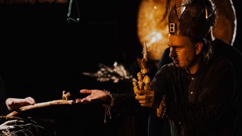  - Zdjęcie ze spektaklu kwiat paproci. Mężczyzna w koronie trzyma w lewej dłoni drewniane figurki ludzi. Prawą dłoń wyciąga ku małej drewnianej figurce kota.