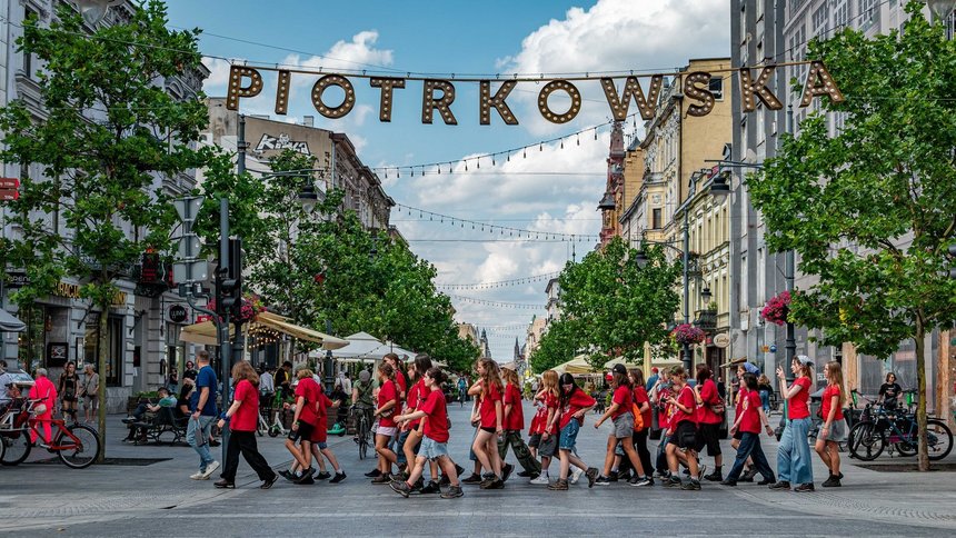 Na zdjęciu grupa młodzieży przechodząca przez ulicę po przejściu dla pieszych. Nad ich głowami na linie zawieszony napis PIOTRKOWSKA.