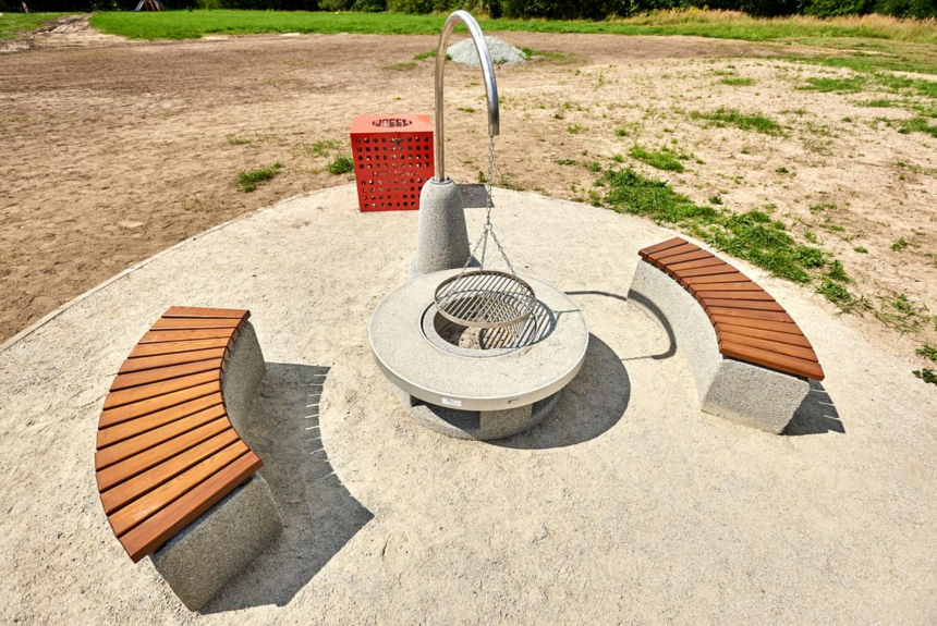 Zdjęcie pokazuje puste miejsce do grillowania - wiszące palenisko na łańcuchu po środku, 2 ławki po bokach oraz kosz na śmieci w Parku na Młynku 