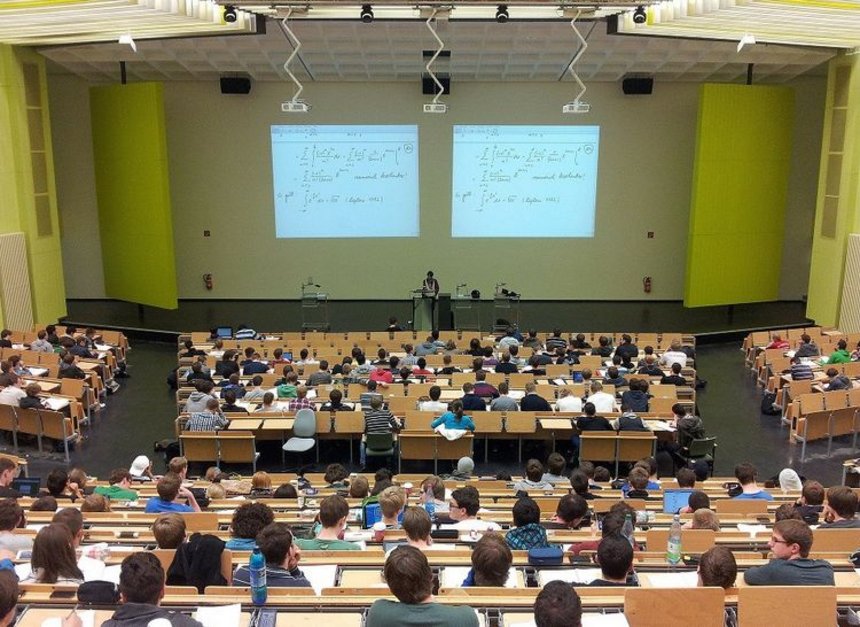 Sala wykładowa na uniwersytecie wypełniona studentami. Za pomocą rzutnika na ścianie wyświetlane są skomplikowane wzory matematyczne.