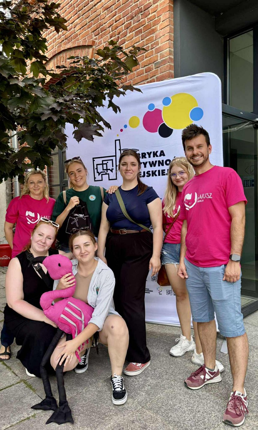 Na zdjęciu widać grupę siedmiu osób w kolorowych koszulka na tle kolorowego banneru reklamowego. Jedna z osób trzyma w ręku maskotkę (różowego flaminga). W tle widzimy fragment korony drzewa na tle fragmentu budynku z czerwonej cegły.