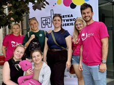 Na zdjęciu widać grupę siedmiu osób w kolorowych koszulka na tle kolorowego banneru reklamowego. Jedna z osób trzyma w ręku maskotkę (różowego flaminga). W tle widzimy fragment korony drzewa na tle fragmentu budynku z czerwonej cegły.