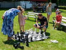 Piknik. Na zdjęciu ludzie grający w wielkowymiarowe szachy na trawie. W tle ławki.