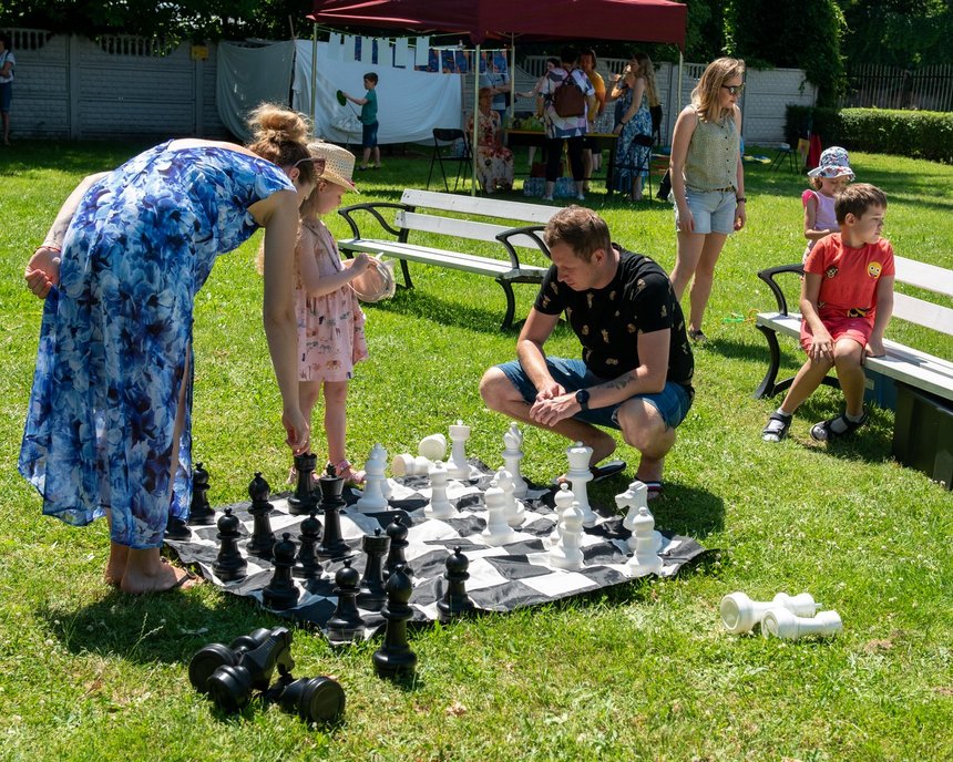 Piknik. Na zdjęciu ludzie grający w wielkowymiarowe szachy na trawie. W tle ławki.
