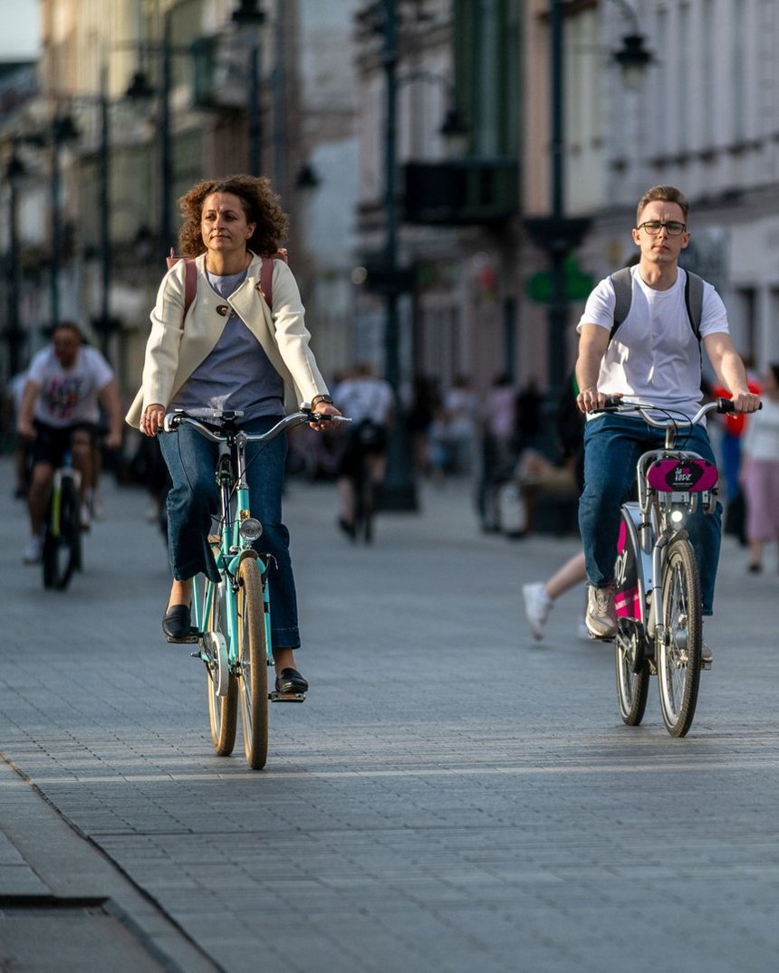 Na zdjęciu na pierwszym planie dwójka ludzi jadących rowerami ulicą Piotrkowską. Kobieta po lewej stronie zdjęcia jedzie na niebieskim rowerze. Mężczyzna po prawej stronie korzysta z roweru miejskiego.