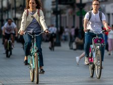 Na zdjęciu na pierwszym planie dwójka ludzi jadących rowerami ulicą Piotrkowską. Kobieta po lewej stronie zdjęcia jedzie na niebieskim rowerze. Mężczyzna po prawej stronie korzysta z roweru miejskiego.