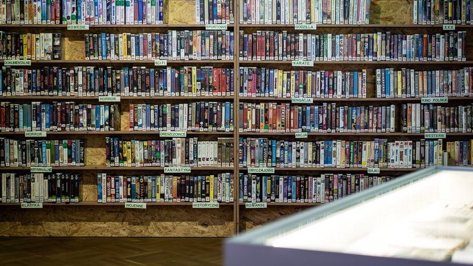  - ściana, na której jest półka z wieloma kolorowymi kasetami video z lat. 90