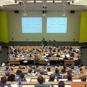 sala wykładowa - aula wypełniona studentami siedzącymi przy długich stołach, w centrum katedra z wykładowcą i wyświetlona na dwóch ekranach ściennych prezentacja