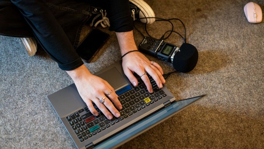 Osoba siedząca po turecku na podłodze, widoczna tylko kawałkiem. Ręce trzyma na klawiaturze laptopa przed sobą. Obok laptopa leży dyktafon z mikrofonem. W prawym górnym rogu rożowa myszka komputerowa.