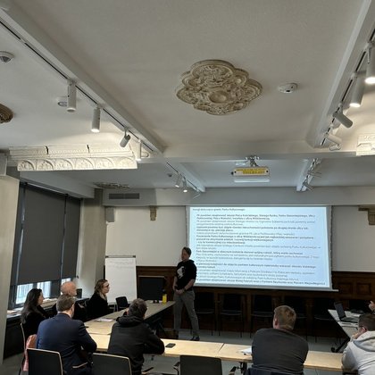 Na zdjęciu widać salę wykładową, gdzie trwa spotkanie. Na ekranie widzimy slajd z tekstem. Uczestnicy siedzą przy ławkach na krzesłach, zwróceni w stronę ekranu. Na zdjęciu widzimy osiem osób. Pomieszczenie jest jasne, po lewej stronie znajdują się zasłonięte okna. Jedna z osób stoi na środku Sali i prawdopodobnie prowadzi spotkanie.