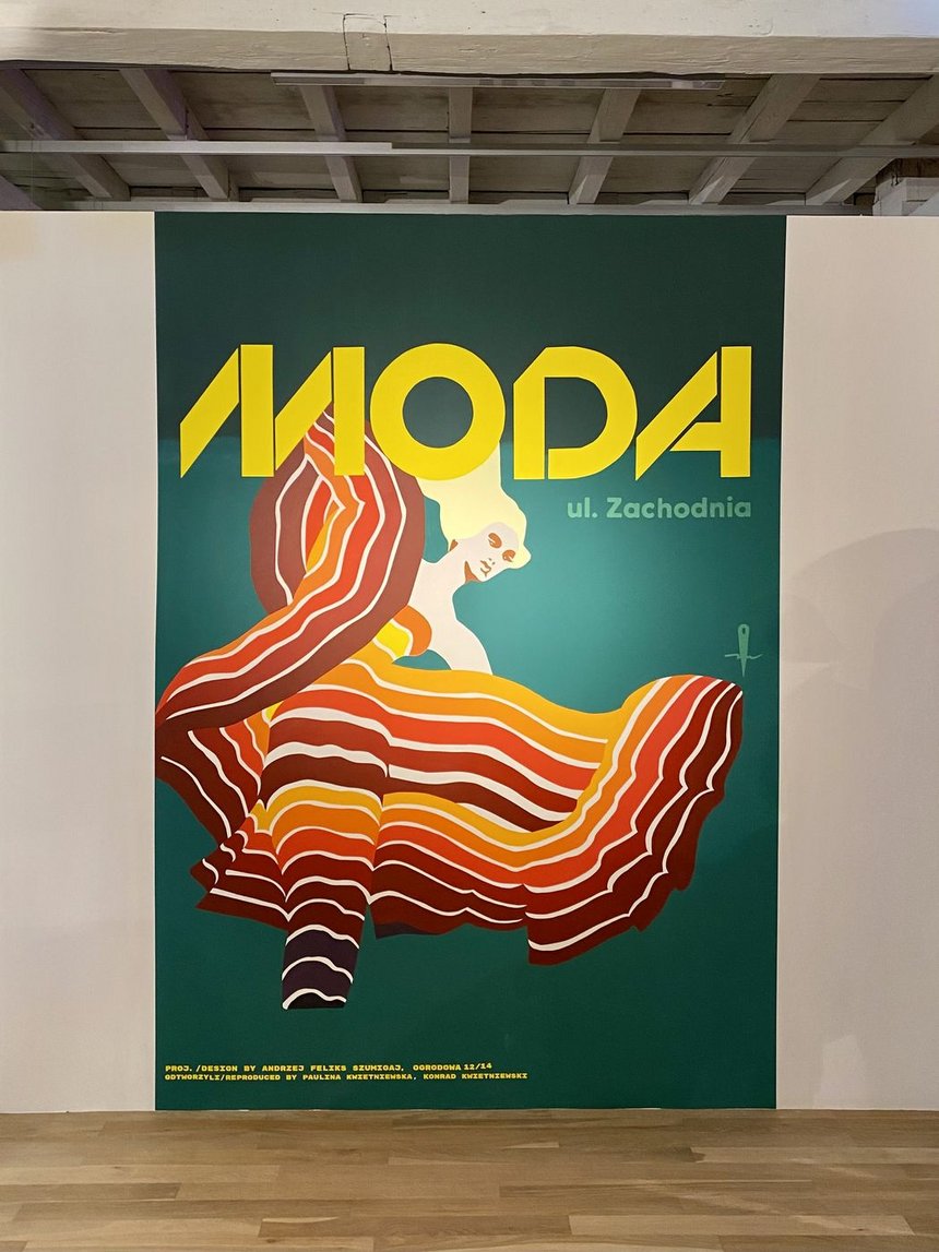 W sali wystawowej wisi wielkoformatowa reprodukcja, a na niej wizerunek kobiet w kolorej sukni. Na górze obrazu napis "MODA".