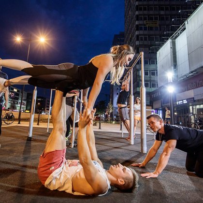 Street work-out. Na pierwszym planie ćwicząca para. Mężczyzna leży na ziemi, podnosi kobietę, którą podtrzymuje uniesionymi kończynami. W tle widać drążki do ćwiczeń i innych ćwiczących.