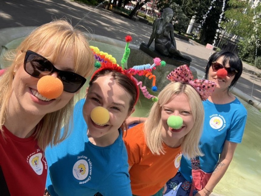 Zdjęcie przedstawia cztery osoby w kolorowych koszulkach i z kolorowymi kulkami na nosach. Zdjęcie zrobione na tle fontanny i rzeźby siedzącej kobiety. W oddali zieleń, drzea i budynki.