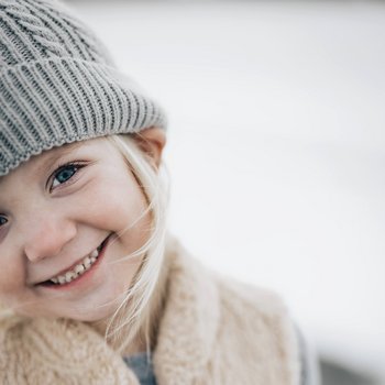 Kilkuletnia dziewczynka o blond włosach wystających spod szarej czapki uśmiecha się ciepło, przechylając lekko główkę. Tło rozmyte.