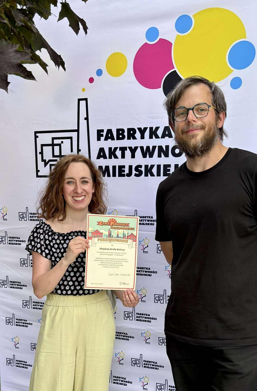 Na zdjęciu widać dwie osoby na tle kolorowego banneru reklamowego z logo fabryki i napisem Fabryka Aktywności Miejskiej. Jedna z osób trzyma w ręku kartkę A-4 z napisami Ziemia Pomagająca Przebudzenie. Jest to podziękowanie dla Miejskiej Strefy Kultury za uczestnictwo w grze miejskiej.