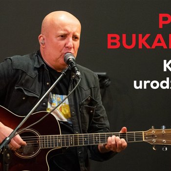 Piotr Bukartyk - mężczyzna w średnim wieku ubrany na czarno, z gitarą w ręku, siedzi na stołku przy mikrofonie.