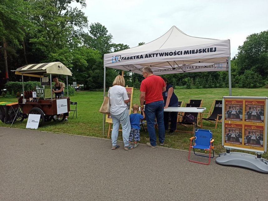 Na pierwszym planie w centralnym punkcie widzimy biały namiot z napisem "FABRYKA AKTYWNOŚCI MIEJSKIEJ" pod namiotem stoi kilka osób dorosłych i jedno dziecko, trzymające osobę dorosłą za rękę. Z prawej strony namiotu stoi niski stojak, w którym umieszczone są cztery plakaty reklamujące portal internetowy "Łódka Wolontariatu" oraz małe rozkładane krzesełko. Po lewej stronie zdjęcia znajduje się niewielki drewniany wózek z daszkiem, przy którym można napić się kawy. Za wózkiem stoi osoba obsługująca. W tle widzimy zieloną trawę i drzewa.