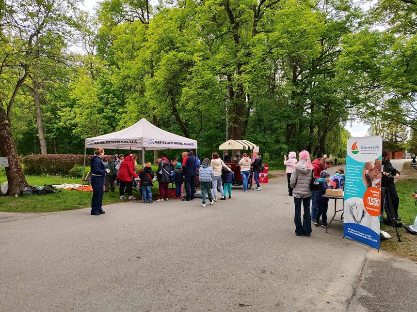 Na zdjęciu widać wydarzenie plenerowe w parku. W centralnym punkcie zdjęcia stoi biały namiot, pod którym zgromadziła się grupa ludzi są tam dorośli jak i dzieci. Obok namiotu z jego prawej stony widzimy daszek wózka gastronomicznego, przy którym stoją dwie osoby. Po prawej stonie zdjęcia stoi mały stolik, przy którym stoi kilka osób i baner reklamowy z napisem Dom w Łodzi. Przy stoliku stoi kilka osób dorosłych i kilkoro dzieci. Jedna z osób robi zdjęcie stoiska gastronomicznego.