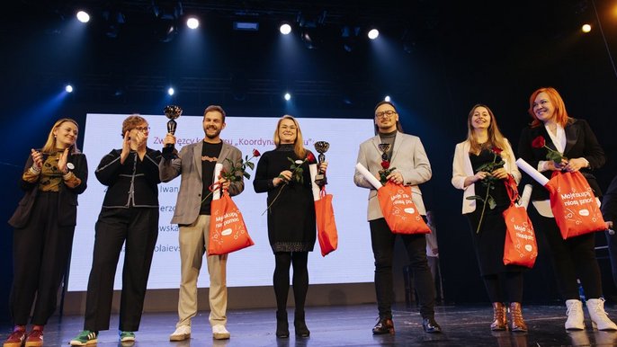 Na zdjęciu widać siedem osób stojących obok siebie na scenie. W tle znajduje się duży biały ekran. Pięć osób trzyma w dłoniach pomarańczowe torby z białym napisem „Moje zakupy niosą pomoc” oraz czerwone róże. Trzy z nich dodatkowo puchar laureata. Uczestnicy są zwróceni przodem do widza i pozują do wspólnego zdjęcia.