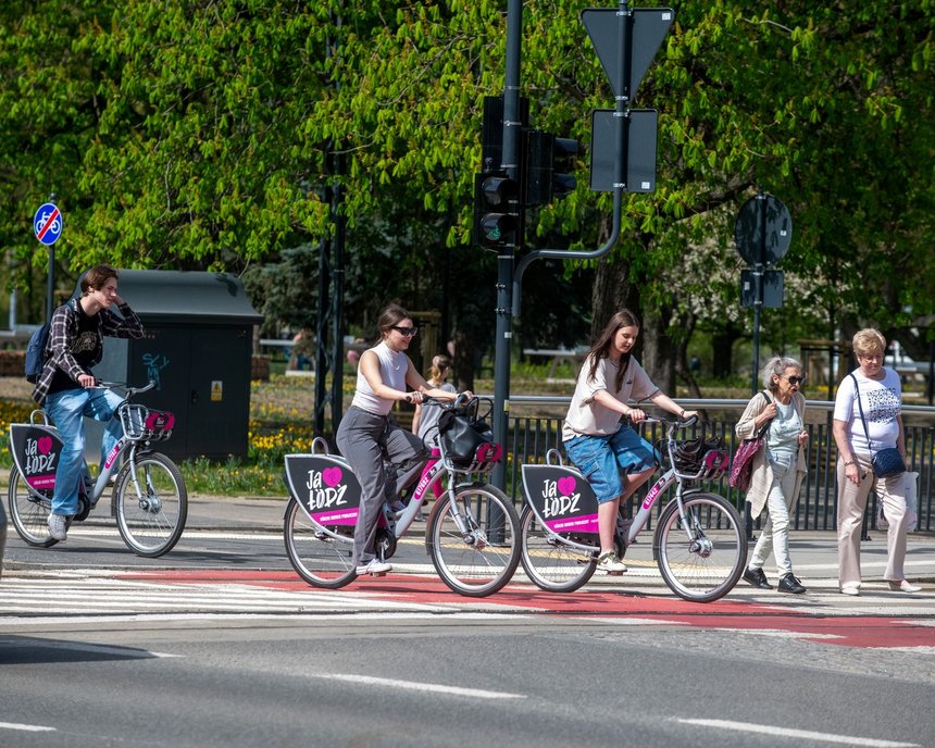 Na zdjęciu ludzie korzystający z rowerów Łódzkiego Roweru Publicznego. Jadą oni wyznaczoną dla rowerów drogą po ulicy. W tle skwer z drzewami.