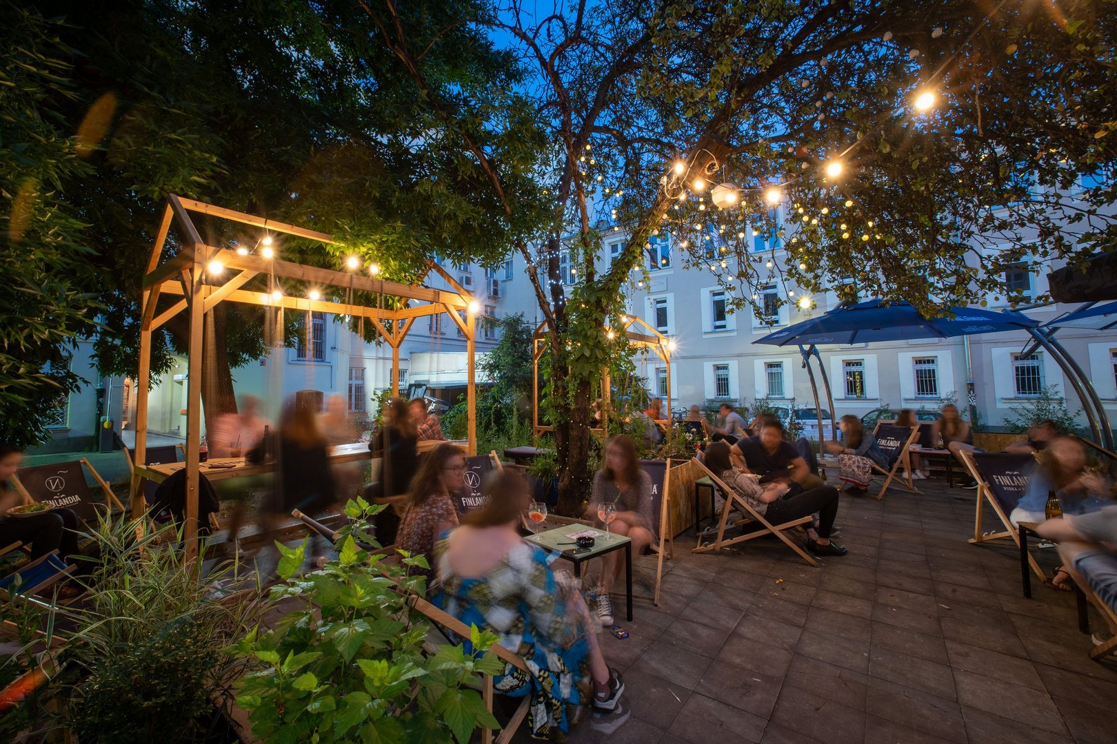 Letni wieczór. Ludzie siedzący na leżakach na dużym podwórku jednej z bram Piotrkowskiej. Na drzewach zawieszone ozdobne lampki w kształcie żarówek.