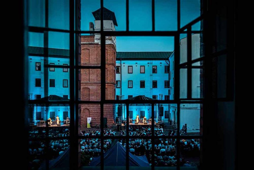 Na pierwszym planie otwarte okno, przez które widać wypełniony ludźmi dziedziniec Białej Fabryki. Na końcu oświetlona scena.