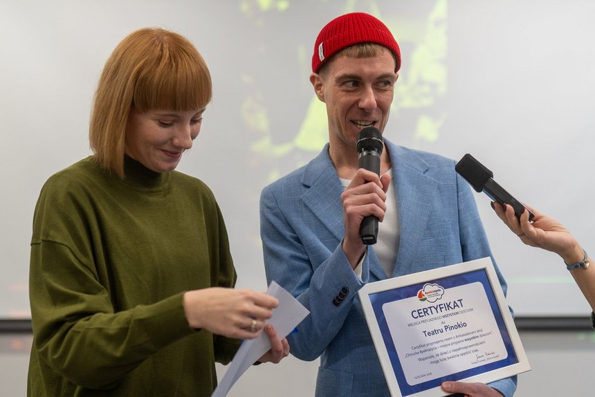 Uśmiechnięty mężczyzna w niebieskiej marynarce i czerwonej czapce na głowie trzyma w lewej ręce certyfikat dla Teatru Pinokio. Mówi do mikrofonu trzymanego w lewej ręce. Po jego stronie stoi rudowłosa kobieta w oliwkowej sukience.