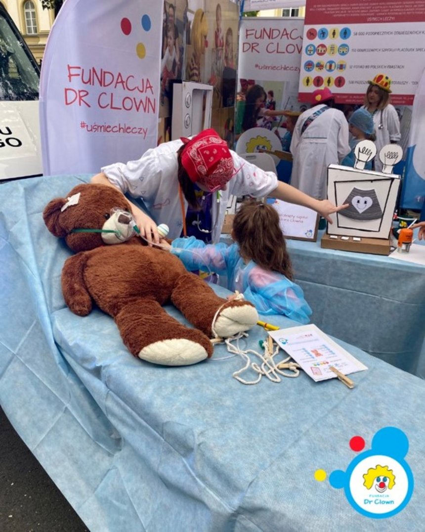 Na zdjęciu widać stoisko Fundacji Dr Clown, organizacji znanej z działań na rzecz uśmiechu i wsparcia emocjonalnego dzieci w szpitalach. Na pierwszym planie znajduje się duży pluszowy miś leżący na niebieskim łóżku szpitalnym – wygląda, jakby był „pacjentem”. Obok niego stoi dziewczynka w niebieskim fartuchu ochronnym, która „bada” misia razem z dorosłą osobą w białym fartuchu, z czerwoną chustką na głowie i kolorowymi elementami stroju. W tle widać banery z logo Fundacji Dr Clown i hasłem „#uśmiechleczy