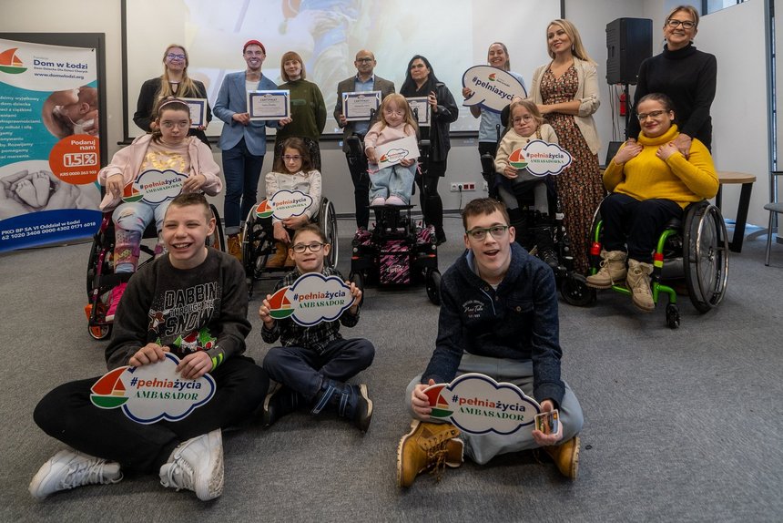 Zdjęcie grupowe. Na pierwszym planie dzieci i młodzież z różnymi niepełnosprawnościami, każde z nich trzyma w rękach kartkę w kształcie chmurki z napisem "#pełniażycia ambasador". Za nimi nagrodzeni z dyplomami w dłoniach.