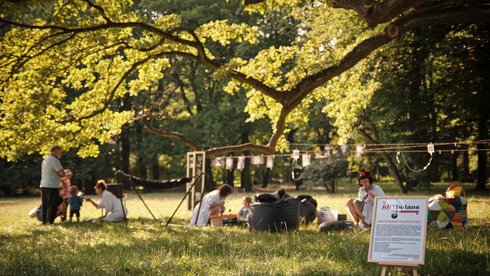 Na zdjęciu zielona polana rozświetlona słońcem. Pod drzewem kilka osób skoncentrowanych w małych grupach. W każdej grupie osoba w białym kitlu.