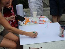 Na obrazku widzimy osobę siedzącą przy stole na zewnątrz. Prawdopodobnie w trakcie jakiegoś wydarzenia lub pikniku. Osoba rysuje coś na dużym białym kartonie zielonym mazakiem. Na stole widzimy kubeczki, kolorowe karteczki i mazaki w przezroczystym pojemniczku. W tle widać nogi innych osób.