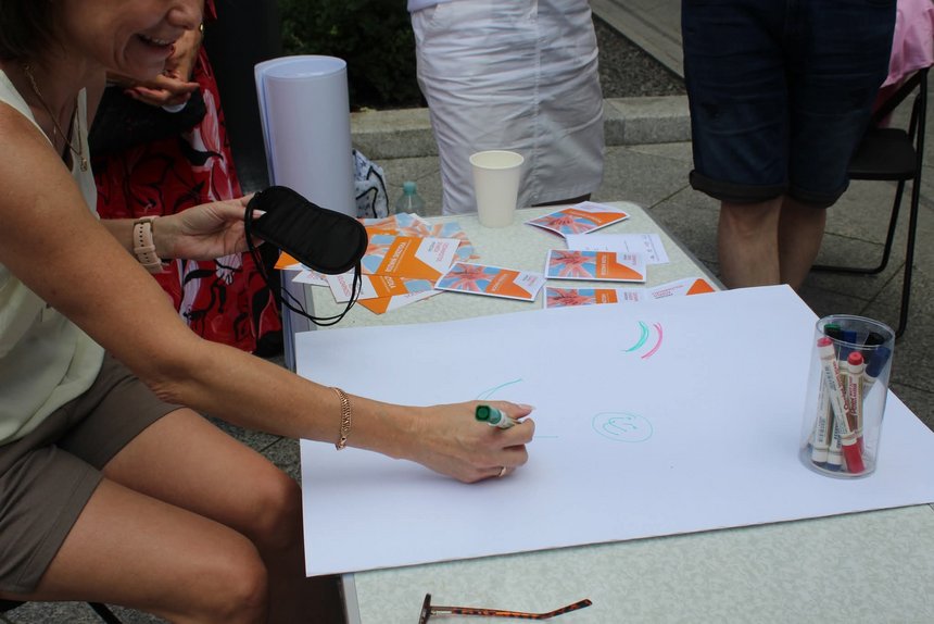 Na obrazku widzimy osobę siedzącą przy stole na zewnątrz. Prawdopodobnie w trakcie jakiegoś wydarzenia lub pikniku. Osoba rysuje coś na dużym białym kartonie zielonym mazakiem. Na stole widzimy kubeczki, kolorowe karteczki i mazaki w przezroczystym pojemniczku. W tle widać nogi innych osób.