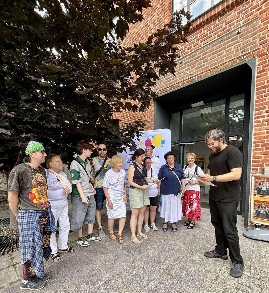Na zdjęciu widać grupę jedenastu osób na tle kolorowego banneru reklamowego z logo fabryki i napisem Fabryka Aktywności Miejskiej. Jedna z osób trzyma w ręku kartkę A-4 w jasnym kolorze. W tle widzimy drzewo a po prawej stronie zdjęcia ścianę kamienicy z czerwonej cegły z widocznym wejściem do budynku. Na dole z lewej strony widać dwa plakaty, na których widać trzy osoby w eleganckich strojach w górnej części plakatu znajduje się napis „Ziemia Pomagająca” a w dolnej, logo Łódki Wolontariatu.