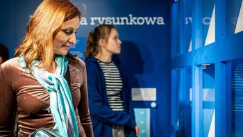  -  Na zdjęciu dwie kobiety. Na pierwszym planie kobieta około czterdziestki w rudych włosach ogląda wystawę. Za nią młoda dziewczyna.